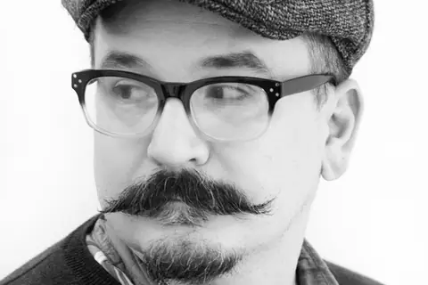 Black-and-white portrait of Jared Pappas-Kelley, wearing glasses and a flat cap.