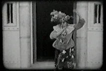 Black-and-white film still showing a figure raising their arms beside an open doorway
