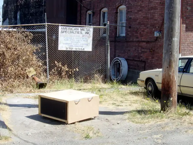 The Story of Home: a mobile wooden audio structure placed in a paved yard beside a fence and brick building
